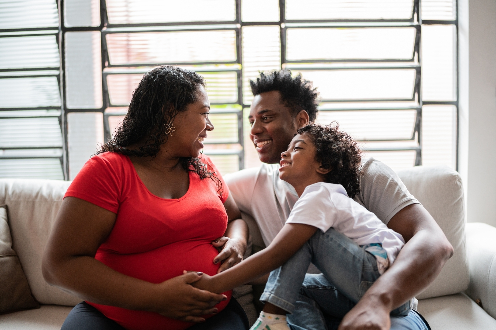 A pregnant mother sits on the sofa at home with her partners and child.