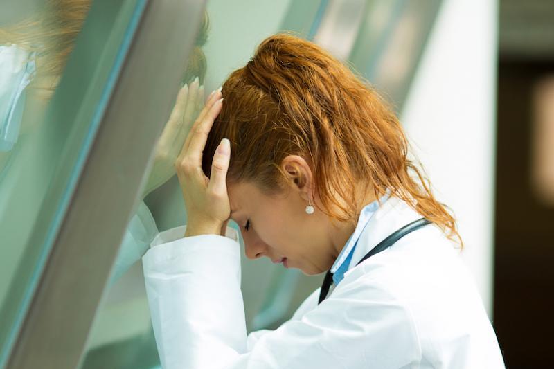 A stressed healthcare worker with hands on forehead, leaning towards a glass wall